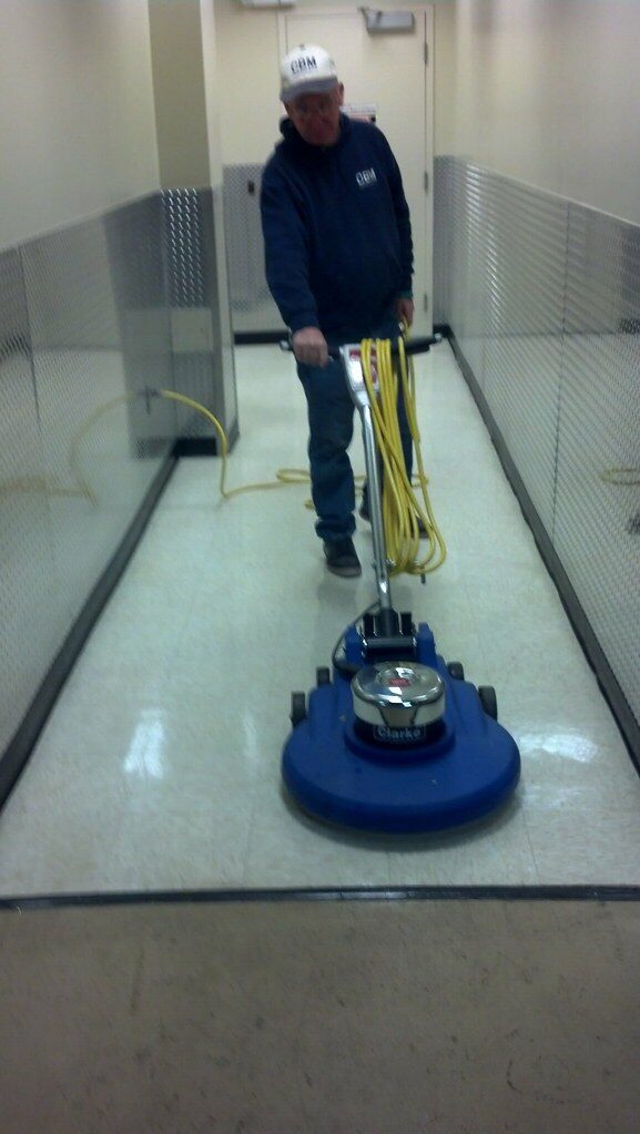 A cleaner in overalls and a cap guiding a rotary floor buffer along a commercial corridor