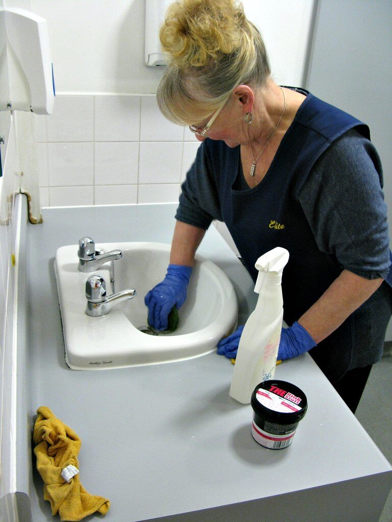 A cleaner in a dark apron and blue gloves scrubbing a white bathroom basin with a spray bottle and cloth