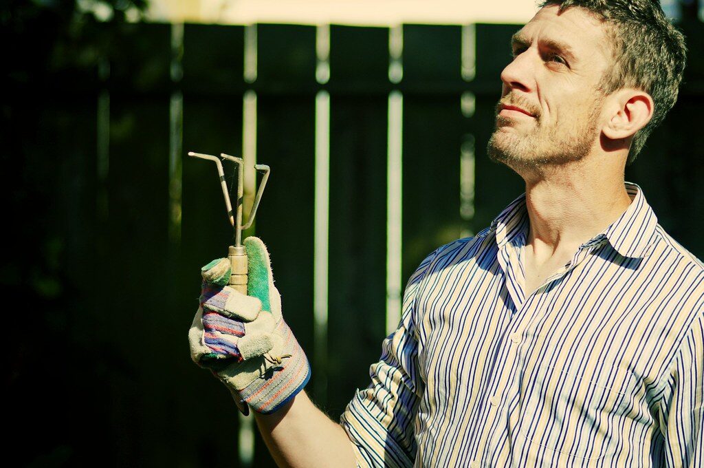 A gardener in work clothes holding pruning shears in front of a dark green hedge