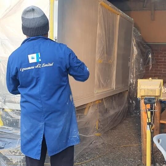 A team member in a blue branded work coat and beanie spray finishing a masked trailer inside a working shed