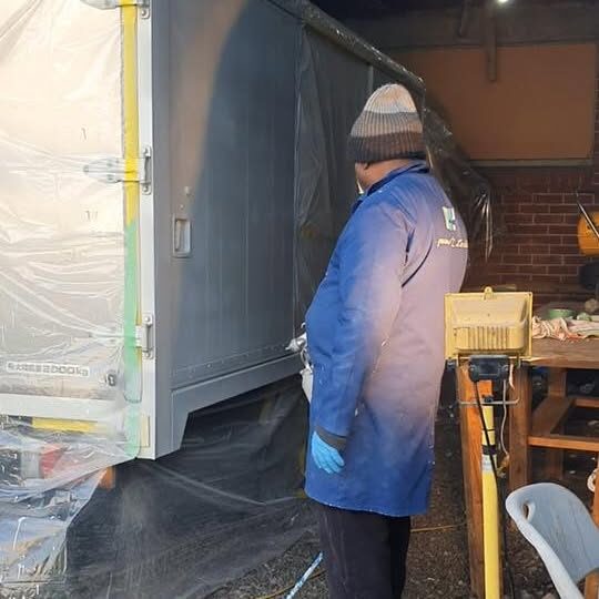 An I Groomers team member in a blue branded work coat spray finishing a trailer panel inside a workshop